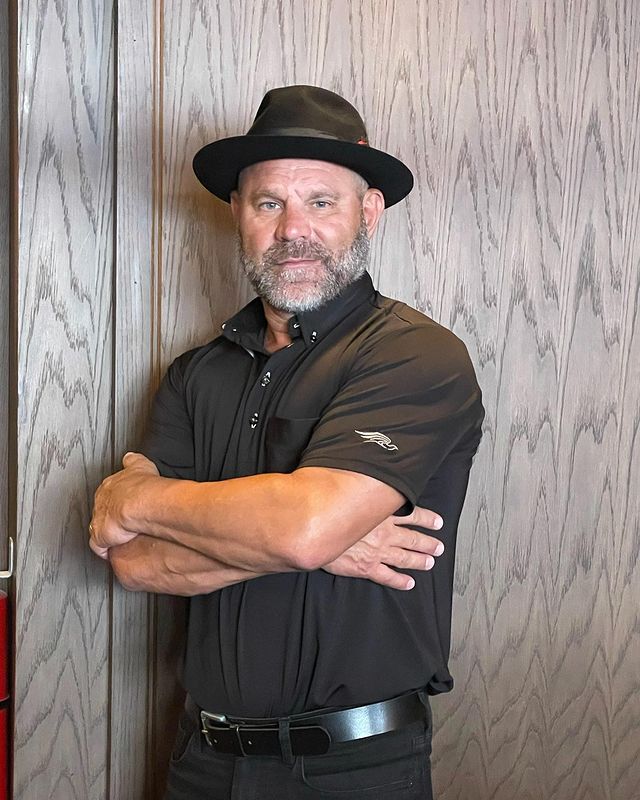 Mark Selman in a black t-shirt, pants, and hat folding his hands.